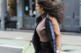 Positive young ethnic female with flowing hair carrying shopping paper bags while quickly crossing road and looking away