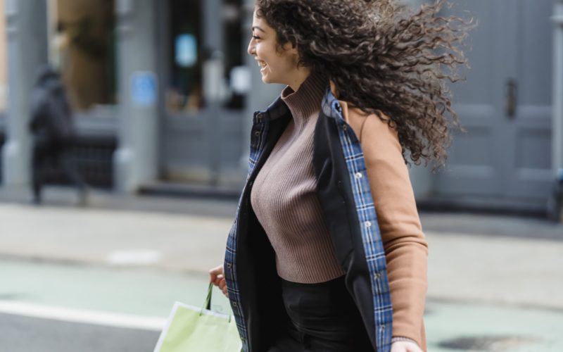 Positive young ethnic female with flowing hair carrying shopping paper bags while quickly crossing road and looking away