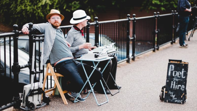 two men sitting on bench while leaning on metal rail near poet hire signboard during daytime